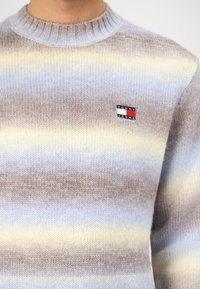 Pull en laine avec des rayures horizontales en marron clair, bleu et jaune. Encolure ronde avec un petit patch logo "Tommy Jeans".
