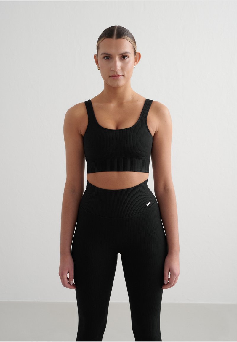 Svart ribbestrikket sports-bh og høydejusterbare leggings med en utskjæringsdesign. Myk tekstur med minimale detaljer på leggingsene.
