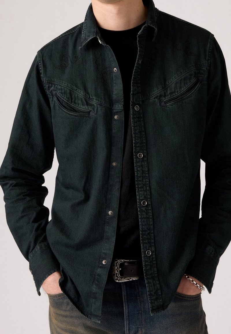Homme portant une chemise en denim boutonnée vert foncé sur un t-shirt noir, associée à un jean foncé, une boucle de ceinture argentée et un bracelet à chaîne.