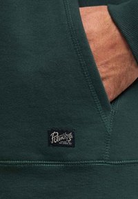 Felpa verde scuro realizzata in tessuto morbido, dotata di tasca. Include un'etichetta blu navy con testo bianco che mostra informazioni sulla marca e sulle cure da adottare.