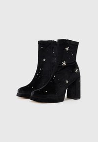 Cizme negre din suede, cu glezna, având un model decorativ cu stele și accente argintii, un toc gros și o închidere cu fermoar pe lateral.