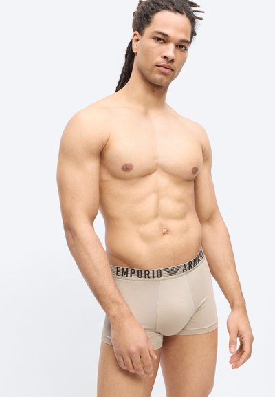 Άνδρας χωρίς πουκάμισο με ντρέντλοκς, φορώντας μπεζ μποξέρ Emporio Armani, στέκεται μπροστά σε απλό ανοιχτό γκρι φόντο.