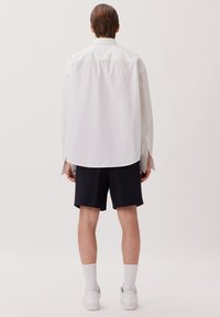 Camicia bianca a maniche lunghe con vestibilità rilassata, abbinata a pantaloni corti blu navy. Il modello indossa sneakers bianche e calze bianche di lunghezza media.