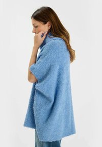 Donna con capelli castani che indossa un ampio poncho-sweater blu a trama, mentre tocca il suo colletto, in piedi di profilo su uno sfondo bianco.