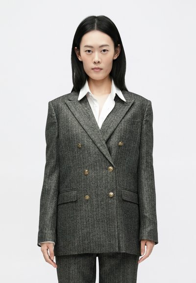 Grijze pinstripe double-breasted blazer met gouden knopen, met uitsparing bij de revers, en twee voorzakken, gedragen over een wit shirt.