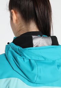 Femme aux cheveux foncés attachés, portant une veste turquoise avec un col à motifs noirs et argentés, vue de dos.