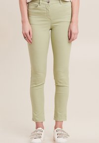 Jeans aderenti verde chiaro dalla texture liscia, design a cinque tasche e chiusura con bottone metallico; abbinati a sneakers bianche con dettagli argentati.