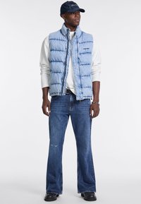 Gilet imbottito blu con motivo cucito, bottoni frontali e colletto alto, indossato sopra una camicia bianca a maniche lunghe e jeans blu scuro. Scarpe nere.