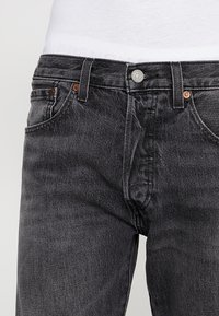 Jeans de mezclilla negra con un acabado ligeramente deslucido. Cuenta con bolsillos delanteros, cierre con botón de metal y acentos de costura marrón.