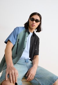 Short-sleeve button-up shirt combining green, black, and blue panels with zippered pockets. Paired with light blue t-shirt and denim shorts.