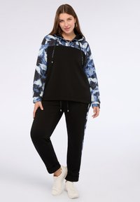 Felpa con cappuccio a pattern nero e blu, con parte anteriore nera unita, cordini regolabili, abbinata a pantaloni neri e sneakers bianche.
