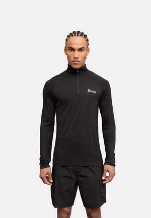 Man met gevlochten haar die een zwart shirt met lange mouwen en een kwart rits draagt met de tekst "Utopia" en zwarte cargoshorts, staand tegen een witte achtergrond.