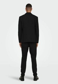 Completo nero con giacca su misura e pantaloni affusolati. Tessuto liscio, due bottoni sulla giacca e design minimalista. Scarpe nere completano l'outfit.