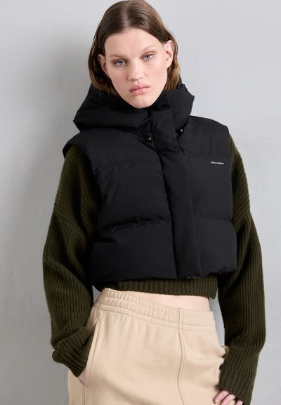 Svart polstret vest med høy krage, med en knappfront og logo. Båret over en mørkegrønn ribbestrikket genser og lysebeige bukser.