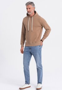 Felpa marrone con cordini regolabili, tasca frontale, abbinata a jeans azzurri chiari e sneakers bianche. Tessuto liscio e vestibilità rilassata.