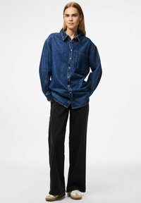 Camicia di jeans blu scuro con bottini e taschino sul petto, abbinata a pantaloni larghi neri e sneakers beige. Scatto in studio su sfondo neutro.