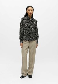 Musta-beige zebrakuvioinen pusero, jossa on solmittava kaula-aukko, yhdistettynä beigen värisiin korkeavyötäröisiin housuihin ja mustiin kenkiin. Sileä, kevyt kangas.