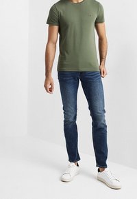 Olivgrünes T-Shirt mit kurzen Ärmeln und Rundhalsausschnitt, versehen mit einem kleinen Logo, kombiniert mit dunkelblauen Slim-Fit-Jeans und weißen Sneakers.