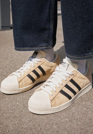 Persona con zapatillas Adidas beige con rayas negras, punteras blancas, cordones blancos, calcetines blancos y vaqueros azul oscuro, de pie sobre hormigón.