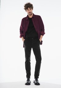 Maroon ruudullinen paita mustan T-paidan päällä; mustat slim-fit -housut; mustat nauhalliset saappaat; sisätiloissa valkoista taustaa vasten.