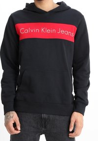 Sudadera negra con una franja roja horizontal que presenta el texto "Calvin Klein Jeans". El material parece suave, con un bolsillo frontal y capucha ajustable.