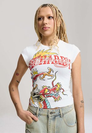 Young person with long blonde braids wearing a white dragon-printed sleeveless shirt and light denim pants, standing with hand in pocket.
