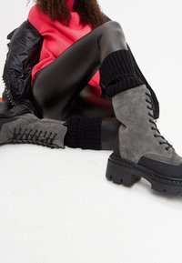 Grijze suède veterschoenen met zwarte rubberen zolen, met een chunky ontwerp en gebreide zwarte boorden. Glanzende zwarte leggings en een roze sweater.