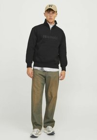 Schwarzer Zip-Hoodie mit dem Schriftzug "ORIGINALS", über einem weißen gestreiften Hemd getragen, kombiniert mit strukturierten beigen Hosen und grauen Turnschuhen.