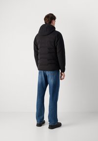 Man in een zwarte gewatteerde hoodie en blauwe jeans, staande met het gezicht naar een effen witte muur met de handen ontspannen naast zich.