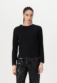 Chemise noire à manches longues avec un col rond, présentant une silhouette ajustée. Associée à un pantalon noir brillant en sequins avec des rayures verticales.