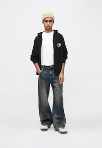 Mlad moški, ki nosi bež kapa, črno pleteno jakno z zadrgo, belo majico, ohlapne izpraznjene kavbojke in bele superge, stoji ob preprostem belem ozadju.