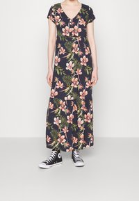 Robe longue avec un fond bleu marine, ornée de grands motifs floraux roses et de feuilles vertes ; manches courtes et décolleté en V, portée avec des baskets noires.