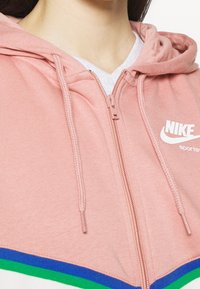 Rosa Nike zip-up hoodie med känguruficka, dragsko och vit logotyp. Har gröna, blå och vita ränder vid fållen.
