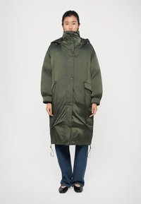 Manteau long matelassé vert avec un col haut, une capuche et des poches avant ; possède une texture brillante, un ourlet ajustable et des épaules tombantes.