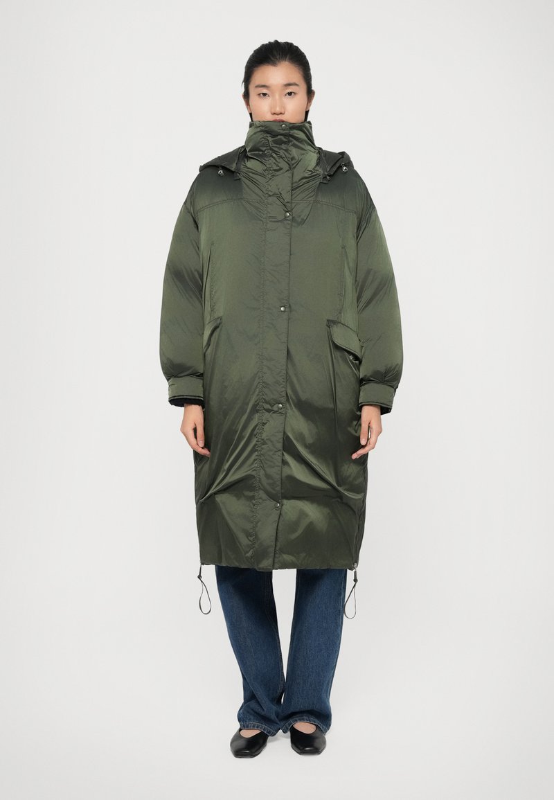 Manteau long matelassé vert avec un col haut, une capuche et des poches avant ; possède une texture brillante, un ourlet ajustable et des épaules tombantes.