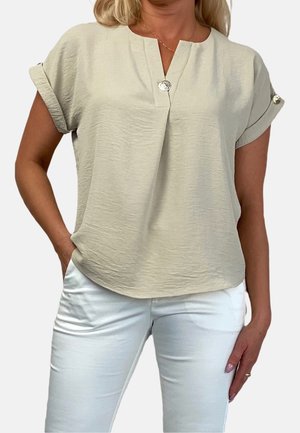 Beige top met korte mouwen, een V-hals en een subtiele plooi. Heeft zilveren knoopaccenten op de mouwen. Gecombineerd met witte broek.