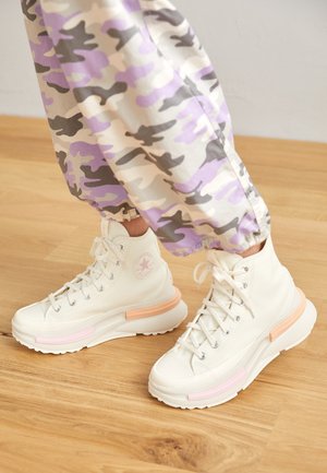 Baskets montantes blanches à plateforme avec des touches roses et couleur bois, portées avec un pantalon camouflage violet clair et gris sur un sol en bois.