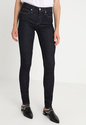 Dopasowane ciemnoniebieskie dżinsy noszone z białym topem i czarnymi, spiczastymi mule ozdobionymi srebrnymi ćwiekami.