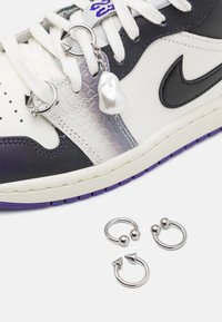 Nike boty s bílým koženým svrškem, fialovými akcenty a vzhledem hadí kůže. Obsahují kovové přívěsky a tři kovové kroužky na bílém pozadí.