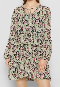 Blumenkleid mit gestuften Schnitt und langen Ärmeln, mit schwarzem Grund, grünen und rosa Blumenmustern sowie einem Bindekragen.