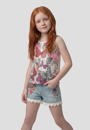 Joven con cabello largo y rojo que lleva una blusa sin mangas con estampado de mariposas y pantalones cortos de mezclilla con bordado floral y ribete de encaje.