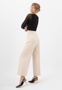 Top lungo a maniche lunghe a coste nera, accorciato, abbinato a pantaloni beige a gamba larga e décolleté neri con tacchi slingback che presentano dettagli dorati. Fondo bianco uniforme.
