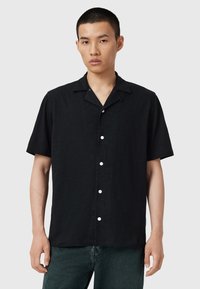 Sort kortærmet skjorte med krave, prydet med hvide knapper og en tekstureret stof, vist på en model mod en neutral baggrund.