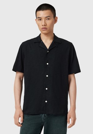 Chemise noire à manches courtes avec un col, dotée de boutons blancs et d'un tissu texturé, présentée sur un mannequin devant un fond neutre.