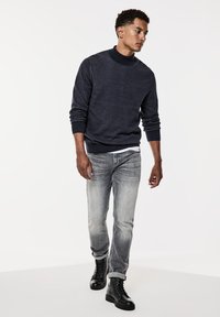 Mörkblå stickad tröja med hög krage, kombinerad med blekta grå jeans och svarta snörstövlar. Har ribbade ärmslut och fåll.