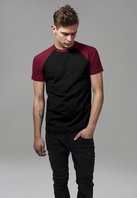 Crna pamučna majica s maroon raglan rukavima. Model stoji s rukama u džepovima, odjeven u crne traperice s rebrastom teksturom.