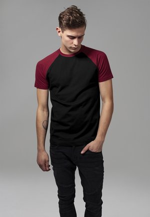 Black cotton t-shirt with maroon raglan sleeves. Model stands with hands in pockets, wearing black jeans with ribbed texture.