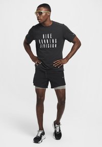 Čierne tričko Nike s nápisom "RUNNING DIVISION" v tučnom bielom písme, kombinované s čiernymi športovými šortkami a čiernymi teniskami. Priehľadné slnečné okuliare so zelenými akcentmi.