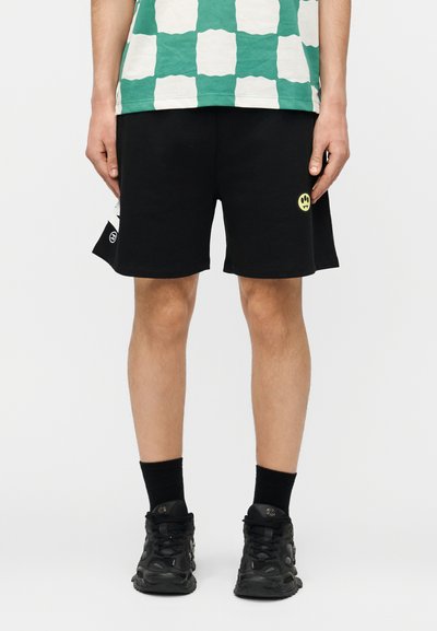 Osoba oblečená v čiernych krátkych nohaviciach s malým žltým logom usmievajúcej sa tváre, zelenej a bielej károvanej košeli, čiernych ponožkách a čiernych teniskách.