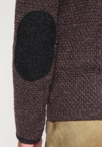 Manche de pull en maille texturée marron avec un grand patch ovale noir au coude, porté par une personne en pantalon beige.
